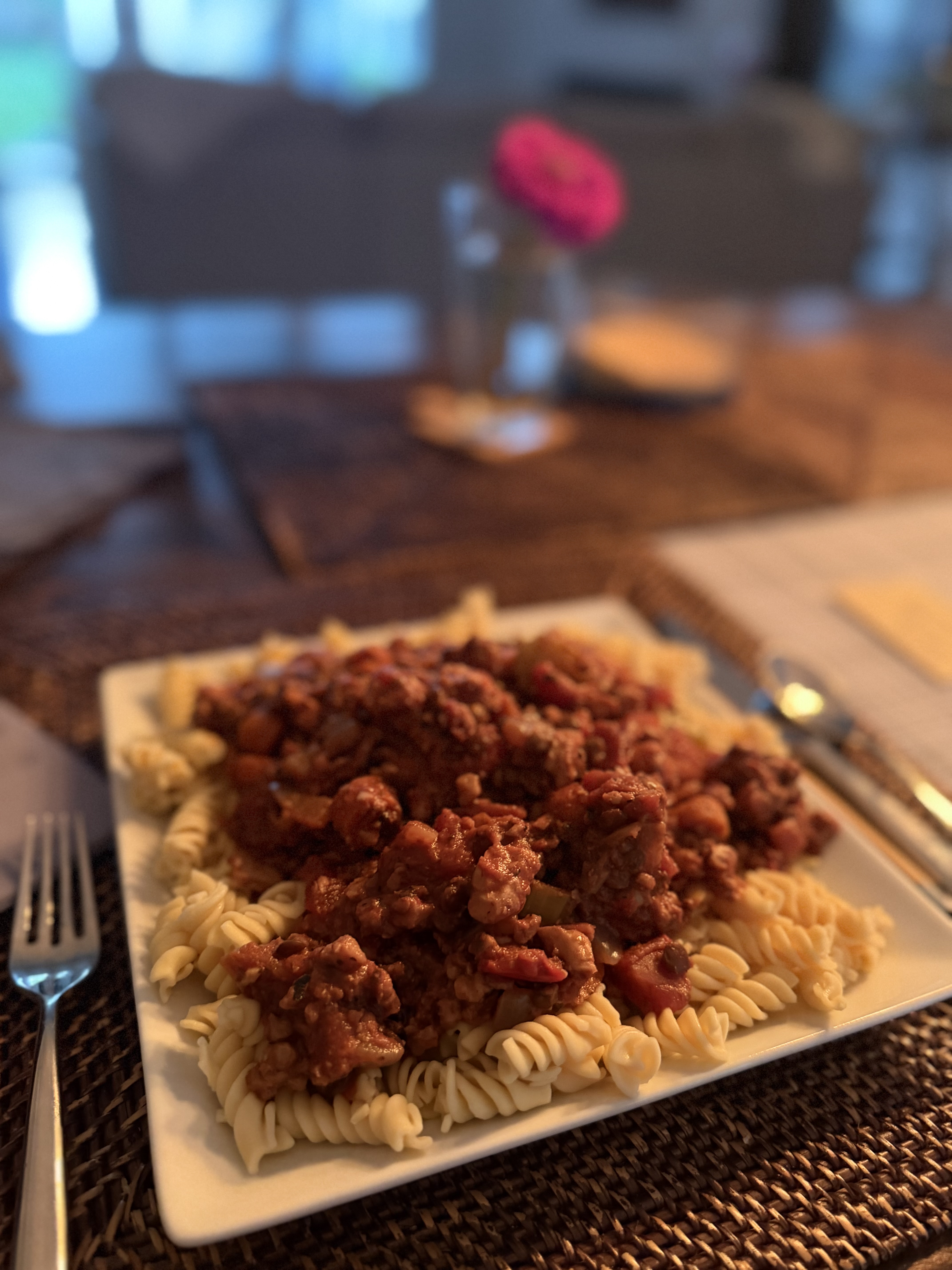 A square white plate of rotini pasta topped with a hearty chickpea and vegetable bolognese sauce