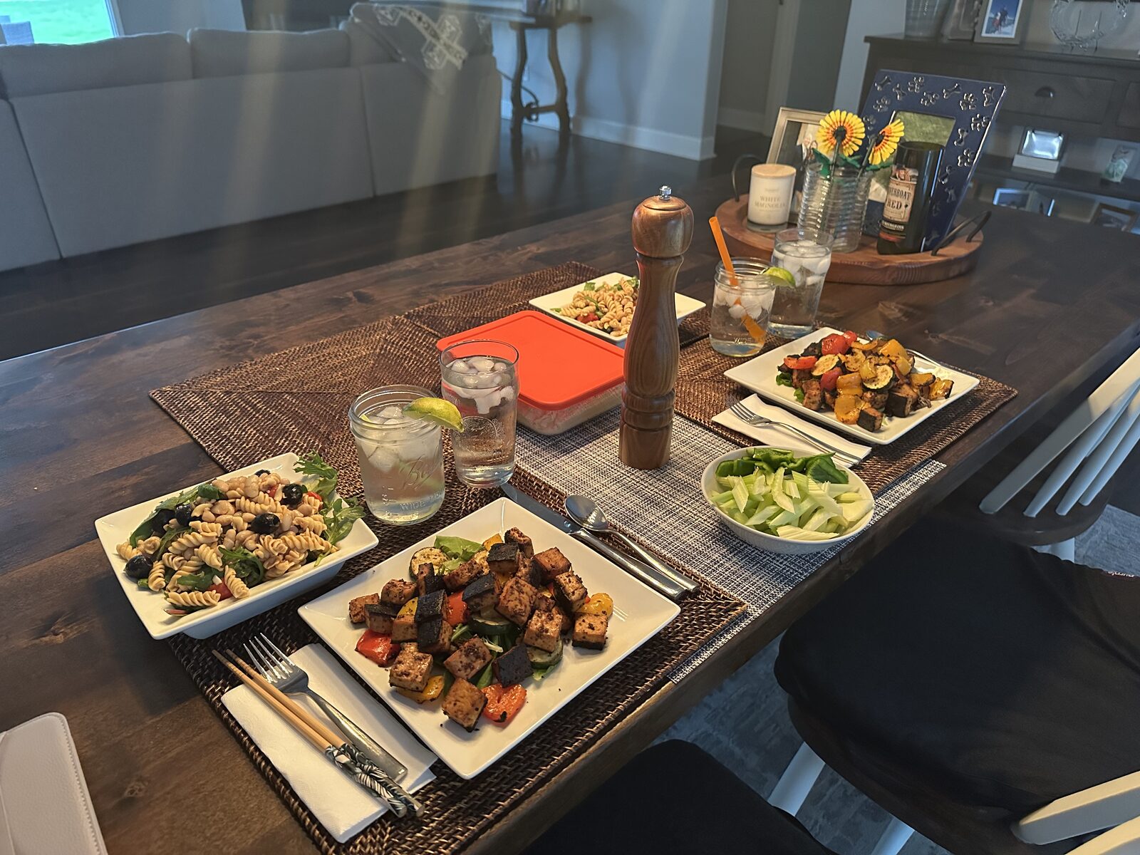 A dinner table set with plates of marinated grilled tofu and charred pepper and zucchini skewers, alongside bowls of white bean pasta salad and sliced celery, with drinks and a pepper grinder
